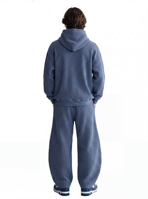 A male model stands in back view with hands in pockets, wearing a muted # blue waffle-knit hooded tracksuit against a plain white background.