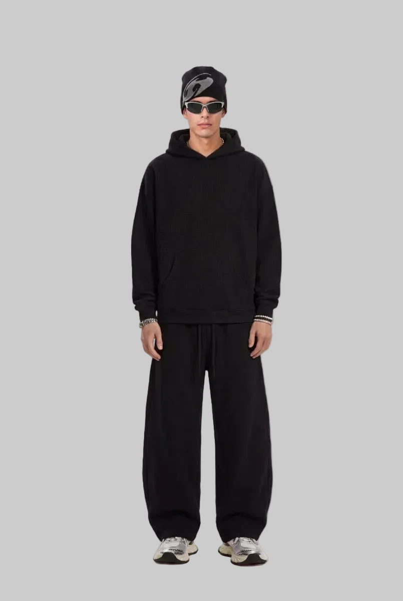 A male model stands facing the camera in a plain white studio setting, wearing a black oversized hoodie, loose #black sweatpants, a black beanie, and silver sneakers. The shot is a full-length fashion look with a clean, minimalist product-photo style.