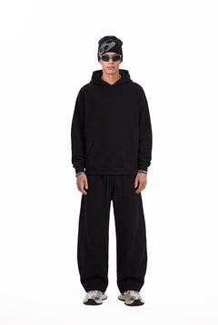 A male model stands facing the camera in a plain white studio setting, wearing a black oversized hoodie, loose #black sweatpants, a black beanie, and silver sneakers. The shot is a full-length fashion look with a clean, minimalist product-photo style.