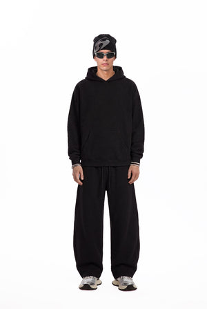 A male model stands facing the camera in a plain white studio setting, wearing a black oversized hoodie, loose #black sweatpants, a black beanie, and silver sneakers. The shot is a full-length fashion look with a clean, minimalist product-photo style.
