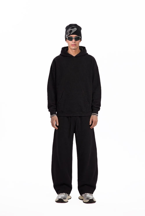 A male model stands facing the camera in a plain white studio setting, wearing a black oversized hoodie, loose #black sweatpants, a black beanie, and silver sneakers. The shot is a full-length fashion look with a clean, minimalist product-photo style.