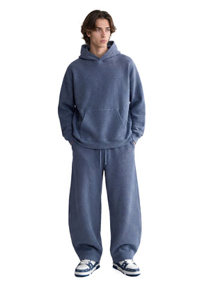A male model stands with hands in pockets against a plain white background, dressed in a muted #blue waffle-knit hoodie and matching wide-leg sweatpants.