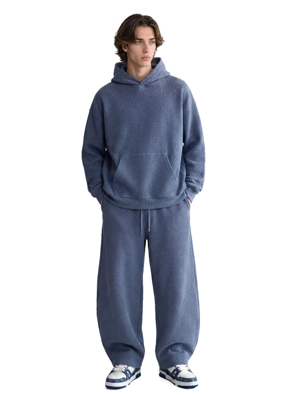 A male model stands with hands in pockets against a plain white background, dressed in a muted #blue waffle-knit hoodie and matching wide-leg sweatpants.