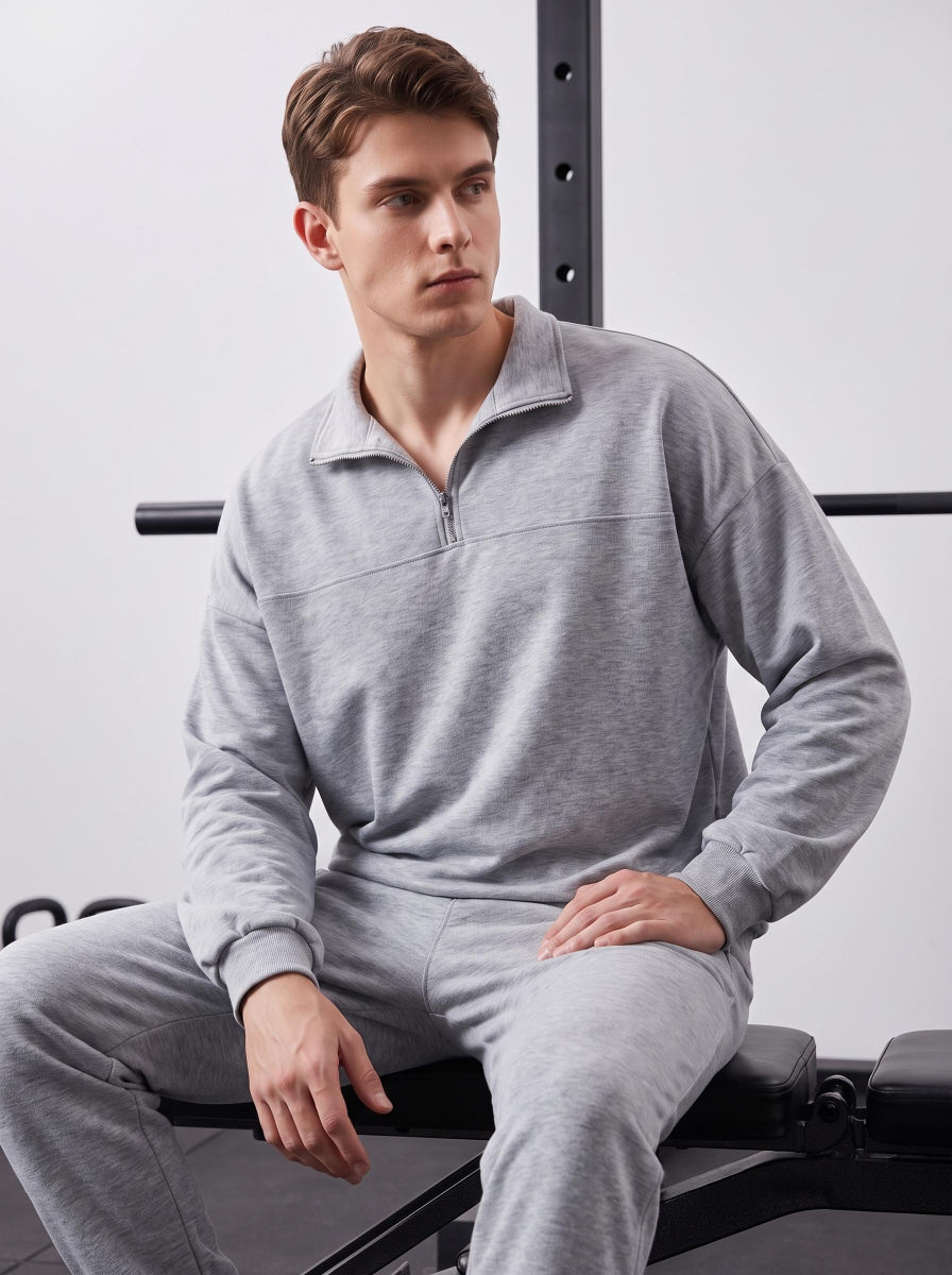 Male model in #grey quarter zip polo matching tracksuit seated on gym bench, gym equipment backdrop, casual sportswear lifestyle shot.