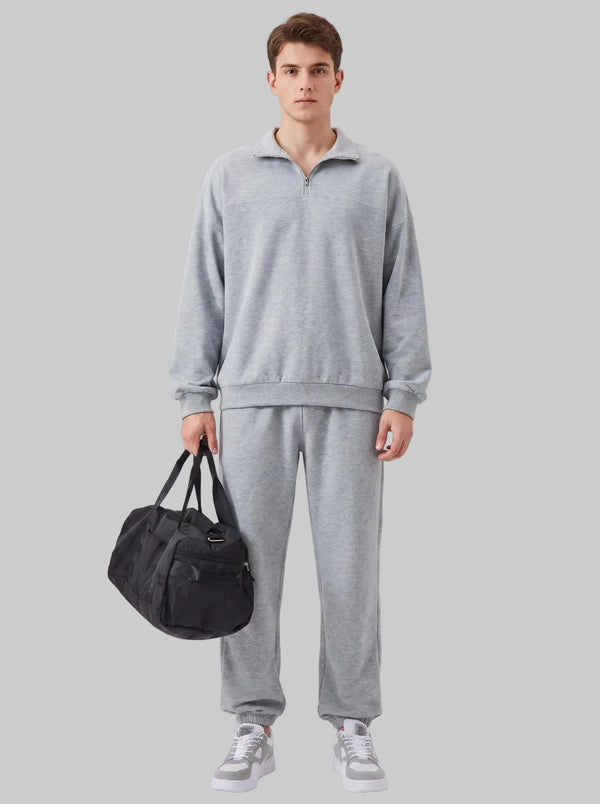 Male model in #grey quarter zip polo tracksuit with joggers, holding black duffel bag, light grey background, casual fashion studio shot.