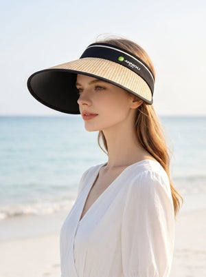 A woman stands on a beach wearing a wide-brimmed sun visor and a white dress, with a calm sea and pale sky in the background.