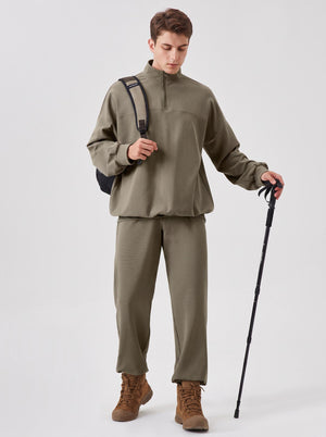 young male model wears an #khaki brown quarter-zip stand collar sweatshirt and matching straight-leg sweatpants. He carries a sling backpack, holds a trekking pole, paired with brown hiking boots. Plain light grey studio background, outdoor casual hiking style,