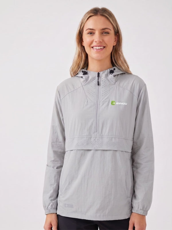 A woman wearing a #lightgray hooded anorak jacket with a half-zip front and a small green and white ANNIOU logo on the left chest, photographed against a plain light gray studio background.