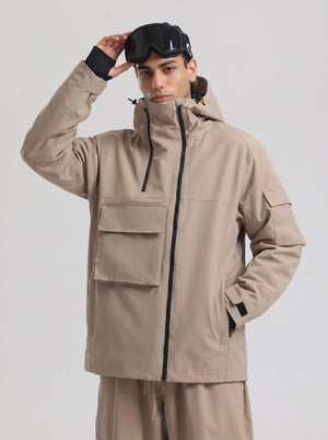 A male model poses against a plain light gray background, wearing a #khaki hooded ski jacket matched with khaki ski trousers. He rests one hand on top of black ski goggles placed on his hood, with the other hand tucked into the jacket side pocket.