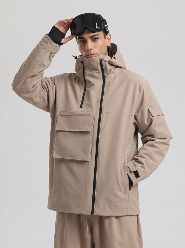 A male model poses against a plain light gray background, wearing a #khaki hooded ski jacket matched with khaki ski trousers. He rests one hand on top of black ski goggles placed on his hood, with the other hand tucked into the jacket side pocket.