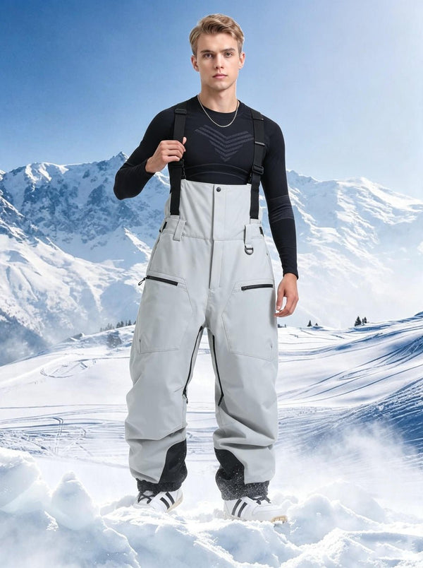 Male model wearing #lightgrey bib ski pants with black suspenders, black thermal base layer top and white‑black ski shoes, standing on snow‑covered ground, snow‑capped mountain range background