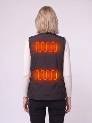 Back‑view shot of a blonde female model wearing a black heated gilet over a white turtleneck top. Bright orange glowing heating‑coil visuals show two heating zones on the vest’s back.