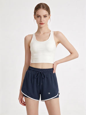 A woman stands in a studio wearing a white cropped athletic tank top and  #navy 2 in 1  running shorts.