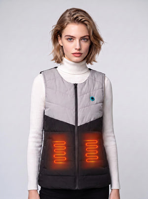 A young European female model wears a two‑tone #greyblack zip‑up heated gilet over a white turtleneck sweater. Glowing orange heating‑coil graphics illustrate the waist heating zones. Plain light‑grey studio background, clean product‑shoot style.