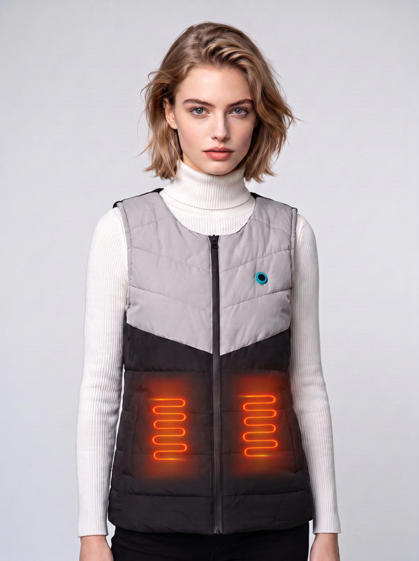 A young European female model wears a two‑tone #greyblack zip‑up heated gilet over a white turtleneck sweater. Glowing orange heating‑coil graphics illustrate the waist heating zones. Plain light‑grey studio background, clean product‑shoot style.