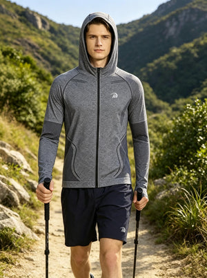 A male model walks forward on a mountain hiking trail, wearing a #grey UPF50+ full‑zip hooded sports jacket with brand logo and breathable mesh panels, paired with navy athletic shorts. He holds two trekking poles. Lush green mountain ranges serve as the natural background under bright daylight. 