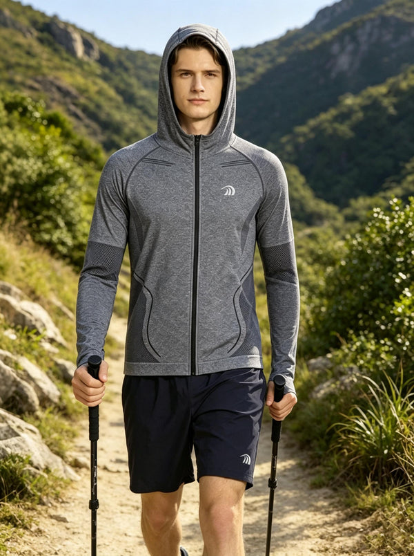 A male model walks forward on a mountain hiking trail, wearing a #grey UPF50+ full‑zip hooded sports jacket with brand logo and breathable mesh panels, paired with navy athletic shorts. He holds two trekking poles. Lush green mountain ranges serve as the natural background under bright daylight. 