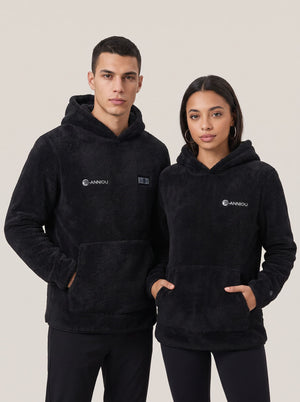 A man and a woman modeling matching black sherpa heated fleece hoodies with a small white 'ANNIOU' logo on the ches