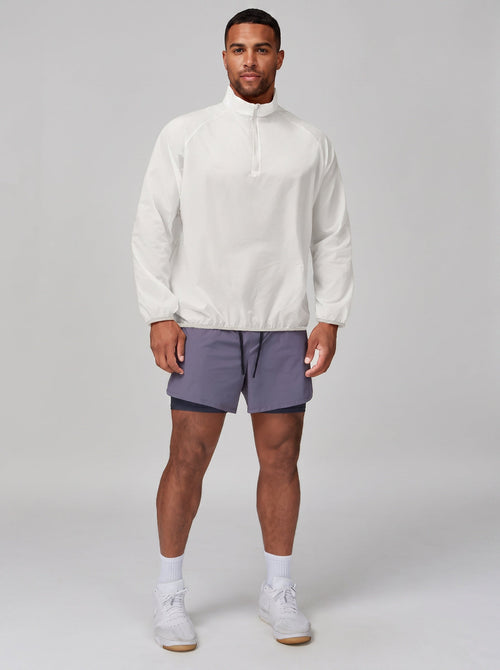 Studio shot against a light grey background. A male model wears a #white half-zip lightweight windbreaker, grey lined athletic shorts, white crew socks and white trainers. 