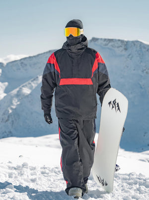 A snowboarder dressed in a black, red and navy color-blocked ski suit walks on thick snow in the mountains. He wears a black beanie, a face covering, and bright orange-tinted ski goggles, holding a white snowboard with black graphic logos in one hand. Snow-covered mountain ranges and a clear pale blue sky form the background.