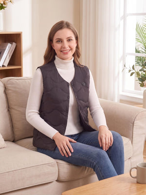 Woman sitting on a beige sofa, wearing a #black snap‑button heated gilet over a cream turtleneck sweater and blue jeans, bright cozy living‑room setting.