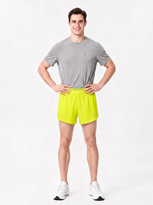 A man stands in a studio wearing a gray short-sleeve athletic T-shirt, bright yellow-green sports shorts.