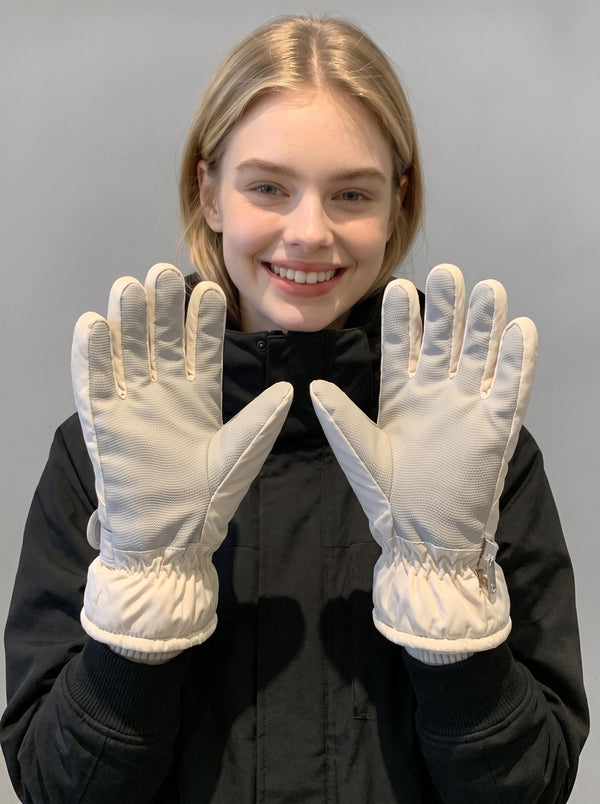 Blonde female model smiles at camera, holding two ski gloves with palms facing forward, showing full‑palm anti‑slip texture, elastic cuffs and side zipper, plain light‑grey studio background.