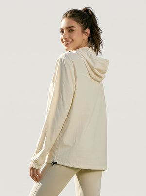 A woman wearing a light cream hooded long-sleeve athletic jacket, shown from a three-quarters back view against a solid light gray studio background.