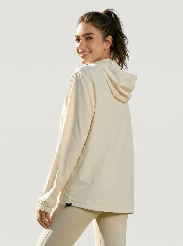 A woman wearing a light cream hooded long-sleeve athletic jacket, shown from a three-quarters back view against a solid light gray studio background.