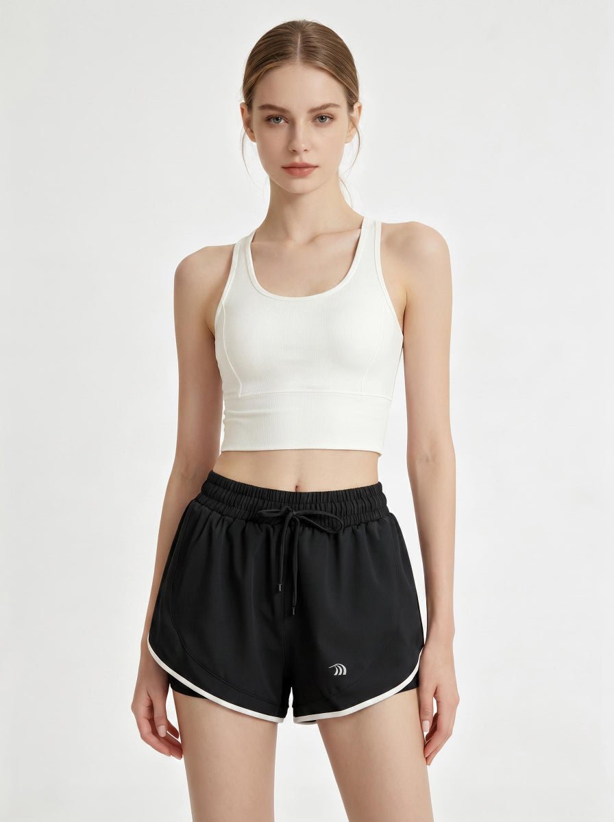 A woman stands in a studio wearing a white cropped athletic tank top and #black 2 in 1  running shorts.