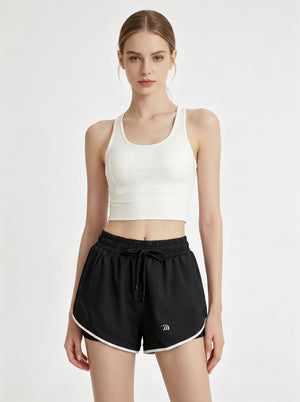 A woman stands in a studio wearing a white cropped athletic tank top and #black 2 in 1  running shorts.