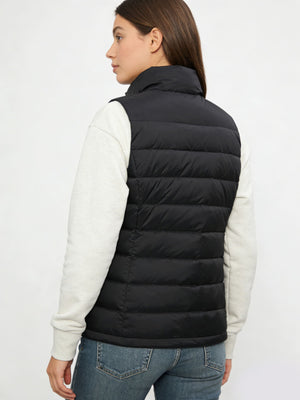 A woman shown from the back wearing a black puffer vest over a light heather gray long-sleeve top and blue jeans, against a plain light gray studio background.