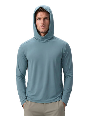 Caucasian male model wearing #blue hooded UPF long-sleeve shirt and khaki trousers, hood up, hands in pockets, plain light gray background.
