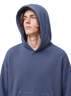 A close-up front view of a male model wearing a muted #blue waffle-knit hoodie with the hood up, set against a plain white background.