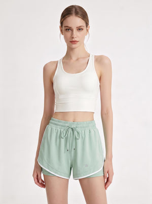 A female model stands in a light gray studio, wearing a white cropped sports tank top and mint-green athletic shorts with white trim, an elastic waistband, a drawstring, and a small logo detail. The image is a clean front-view product shot focused on activewear.