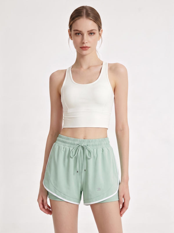A female model stands in a light gray studio, wearing a white cropped sports tank top and mint-green athletic shorts with white trim, an elastic waistband, a drawstring, and a small logo detail. The image is a clean front-view product shot focused on activewear.