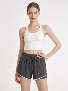 A woman stands in a studio wearing a white cropped athletic tank top and #grey 2 in 1  running shorts.
