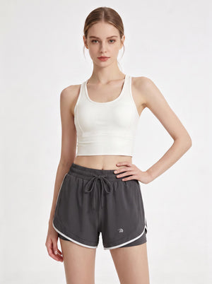 A woman stands in a studio wearing a white cropped athletic tank top and #grey 2 in 1  running shorts.