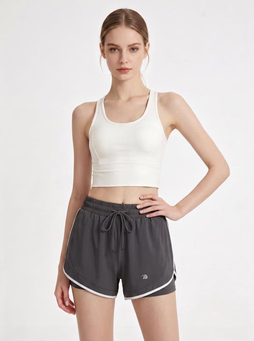 A woman stands in a studio wearing a white cropped athletic tank top and #grey 2 in 1  running shorts.