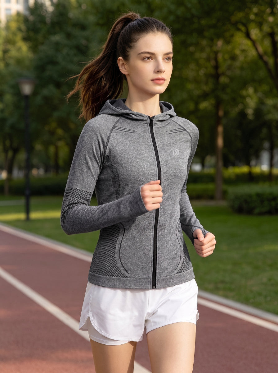 A female model jogging on an outdoor running track, wearing a #grey zip‑up hooded athletic jacket with mesh breathable panels and thumb‑hole cuffs, paired with white sports shorts. 
