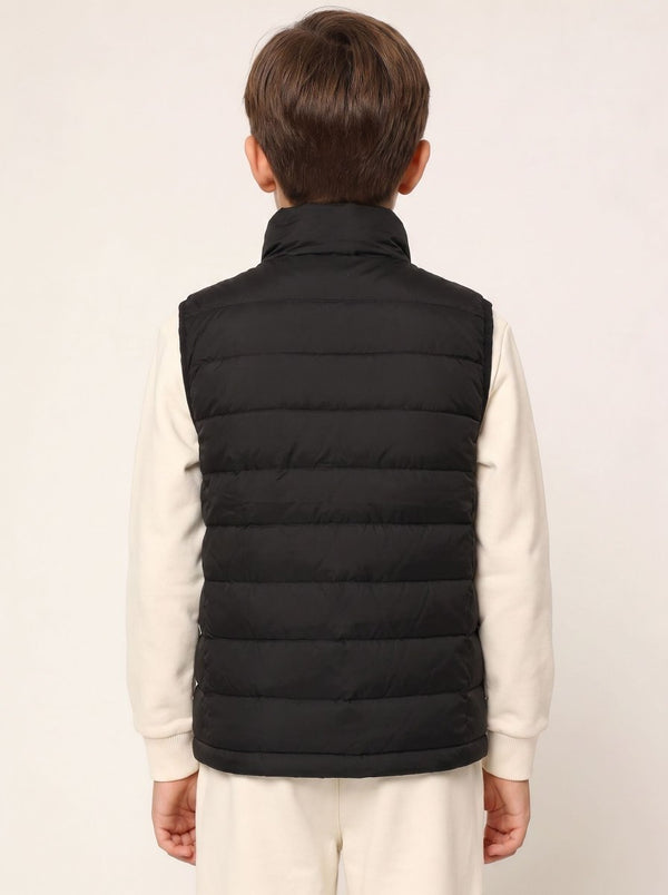 A child viewed from the back wearing a black quilted puffer vest over a cream-colored long-sleeve shirt and cream pants, against a plain off-white studio background.