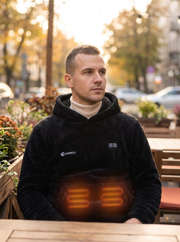 A man sitting at an outdoor cafe table wearing a black fuzzy fleece hoodie with a glowing orange heated seat pad underneath. The background shows a warm, blurred street scene with autumn trees.