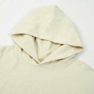 A close-up shot of a cream waffle-knit hoodie laid flat, showing the hood texture and stitching against a plain white background.