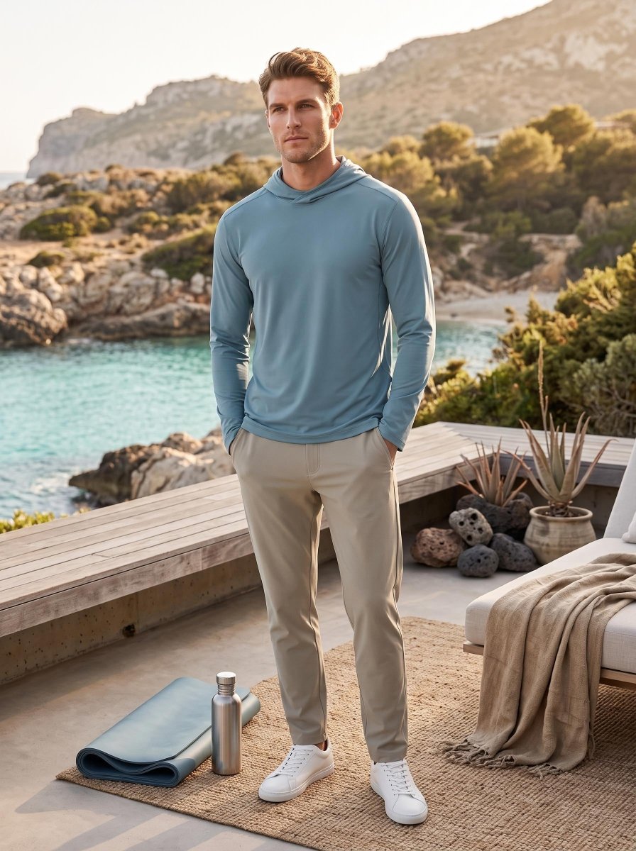 A male model in a #blue hooded long-sleeve top and khaki trousers stands on a seaside terrace. The scenic coastal backdrop features turquoise water and rocky hills. A yoga mat and water bottle rest nearby, creating a relaxed premium outdoor lifestyle scene.