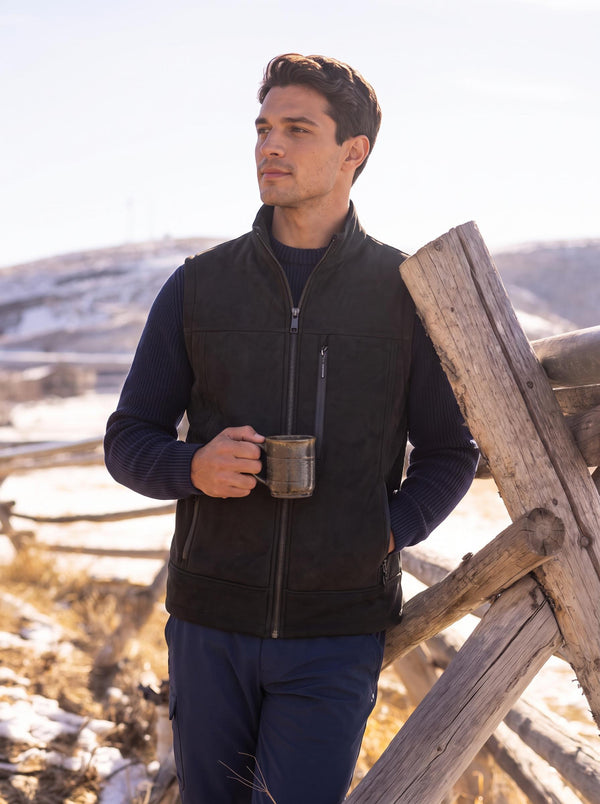 Handsome man in black zip‑up stand‑collar vest over navy knit sweater, holding metal mug, leaning against weathered wooden fence, wintry rural hills background with patches of snow under bright daylight.