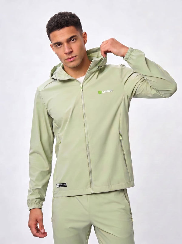A man wearing a #lightgreen hooded jacket and matching pants, posing against a light gray studio background. The jacket features a small green and white logo Anniou on the left chest and a black brand patch near the hem.