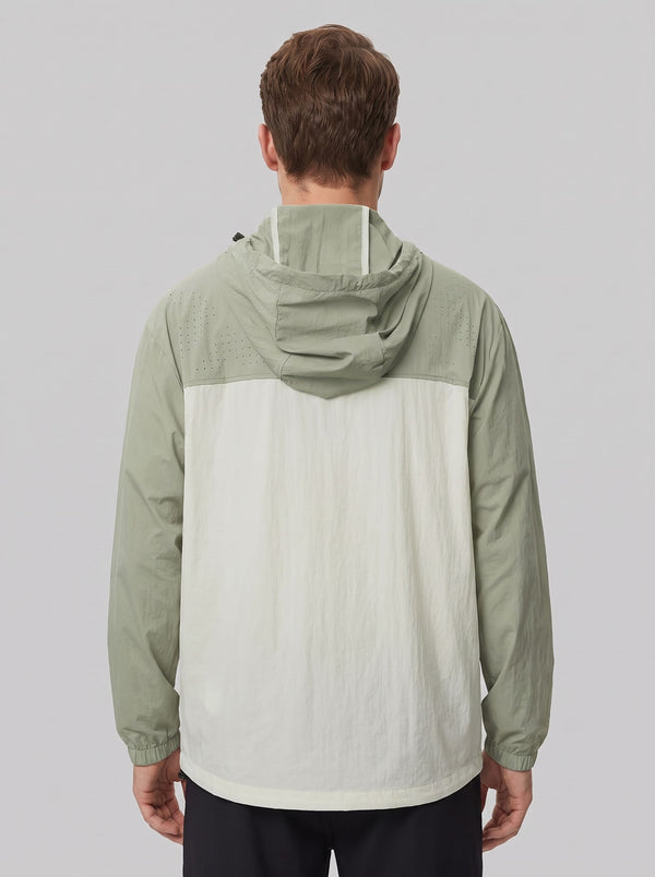 A man viewed from the back wearing a lightweight hooded jacket with a sage green hood and shoulders and an off-white body, set against a solid gray studio background.