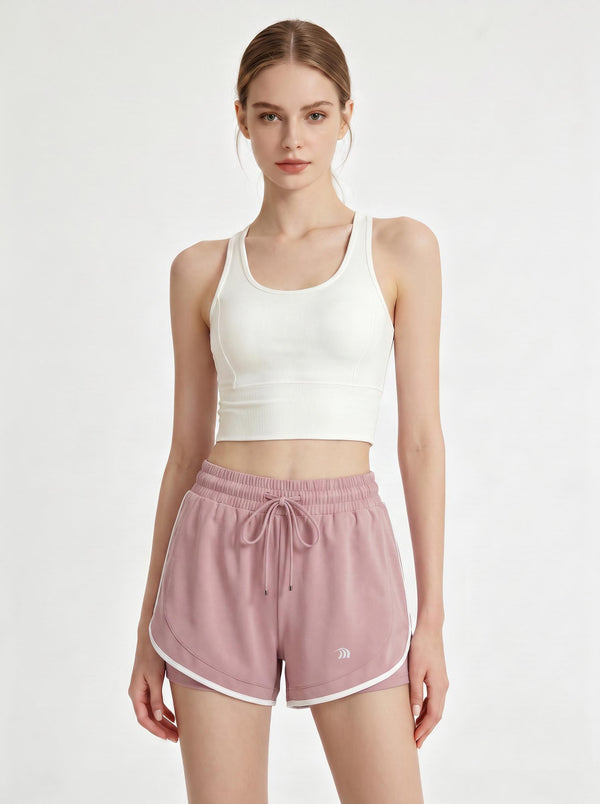 A woman stands in a studio wearing a white cropped athletic tank top and #pink 2 in 1  running shorts.