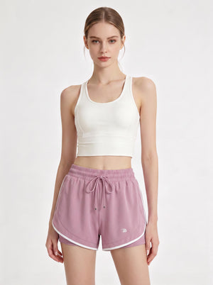 A woman stands in a studio wearing a white cropped athletic tank top and #purple 2 in 1  running shorts.
