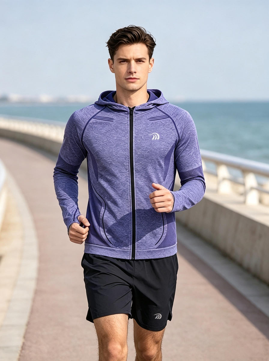 A male model jogging on a seaside coastal promenade, wearing a #purple full‑zip hooded sports jacket with brand logo, mesh breathable panels on sleeves and sides, paired with black athletic shorts. The ocean and soft‑focus seaside railing form the background, bright natural daylight.